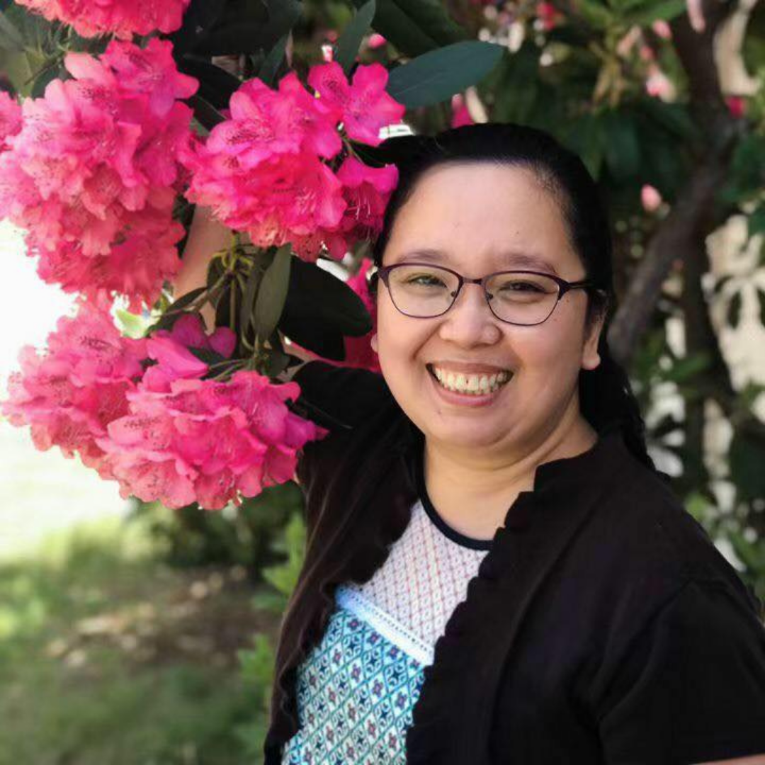 A smiling woman wearing a blue and black top, posing next to pink flowers. 